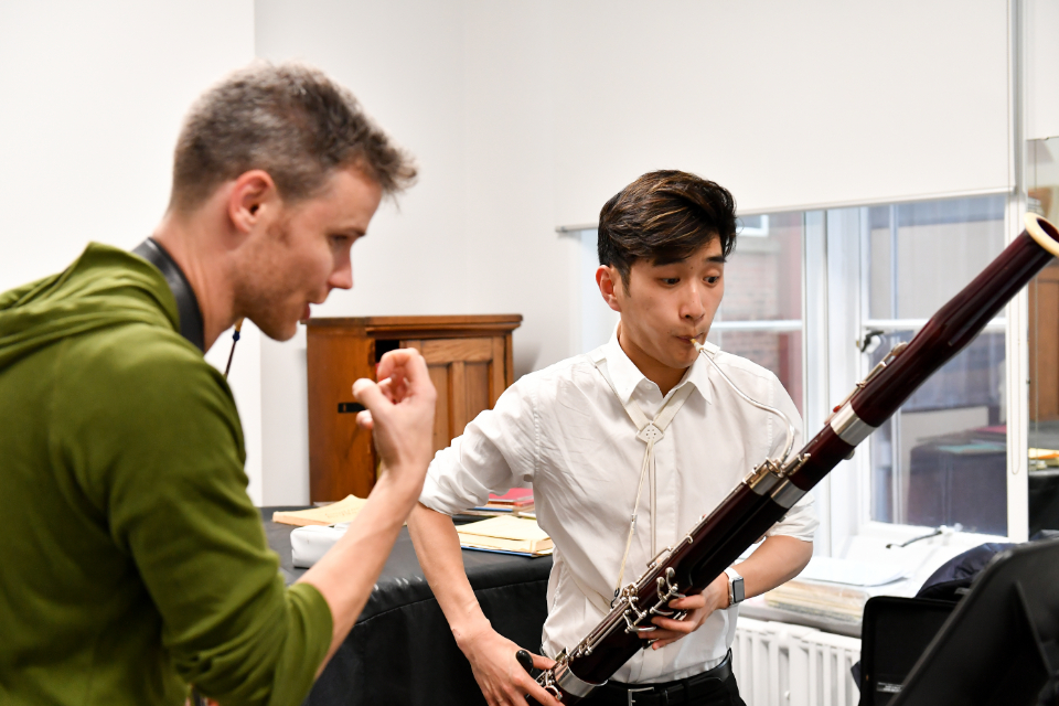 A male professor helping teach a male student, who is performing on a bassoon, in a one to one lesson.